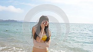 Young attractive woman in a swimsuit is talking on mobile phone on a sea beach. Serious girl in bikini speaking on the