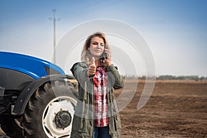 A young attractive woman is talking on the phone in front of a tractor in a field