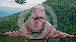 A young attractive woman traveler with a backpack in casual clothes and a pink hoodie stands on top of a mountain. A