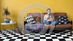 Young attractive woman types emotional message on her phone. Stock footage. Young woman sits on couch and quickly types