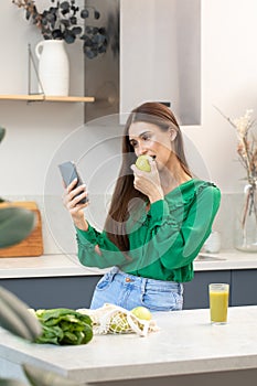 A young attractive woman uses the phone while standing in the kitchen and eating a green apple.