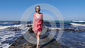 Young attractive woman walking barefoot on the sea pier towards the camera in red dress and sunglasses. Stormy waves hitting the p