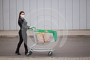 Young attractive woman walking with trolley groceries from a supermarket. Social distancing for protection, face mask to