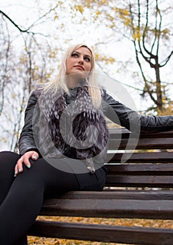 A young attractive woman wearing fur black jacket holding book sitting on the bench in falling park.