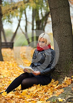 A young attractive woman wearing fur black jacket holding book sitting by the tree. Walking around autumn park.