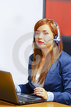 Young attractive woman wearing office clothes and headset sitting by desk looking at computer screen, working with