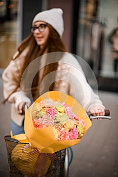 Young attractive woman wearing spring or autumn outfit holding bicycle with multicolored carnation flowers bouquet in