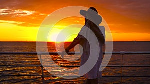 Young attractive woman wearing white dress and hat posing on sea background at amazing sunset