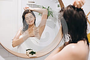 Young attractive woman with wet hair in white towel enjoying smooth skin after shaving armpits, reflection in mirror in stylish