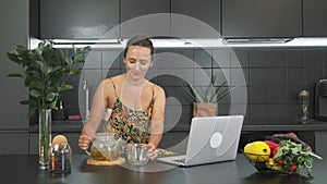 Young attractive woman is working on laptop computer at home in modern kitchen. Thoughtful smiling girl is drinking herbal tea on