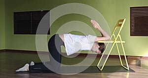 Young attractive woman working out in sportswear with a chair, performing an exercise to improve the mobility of shoulder joints