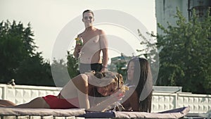 Young attrective man speaks with two shy and smiling girls, drinking coctails on the lounge near swimming pool