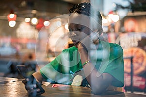 Young authentic black woman sitting with phone in city coffee shop at night