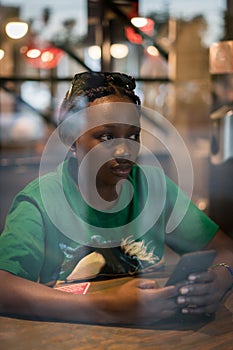 Young authentic black woman sitting with phone in city coffee shop at night