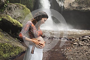 Young authentic pregnant woman near amazing waterfall. Harmony with nature