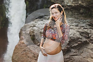 Young authentic pregnant woman near amazing waterfall. Harmony with nature