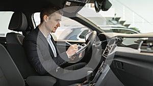 Young auto salesman sitting inside brand new car, making checkup, writing down data at modern dealership, panorama