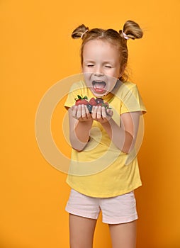 Young baby girl kid with a handful of berries fruits in her palms hands screaming with happiness or in fright on yellow