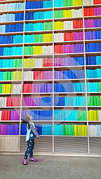 A young baby girl in the library standing near an endless bookshelf with colourful row of books.