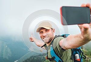 Young backpacker man taking selfie picture using smartphone and showing Thumbs Up during walking by the foggy cloudy weather