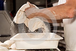 Young baker in bakery shop sprinkling flour with strainer on fresh bread dough in front of oven. concept of traditional manual