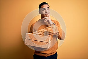 Young baker brazilian man holding box with homemade bread over isolated yellow background serious face thinking about question,