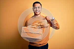 Young baker brazilian man holding box with homemade bread over isolated yellow background with surprise face pointing finger to