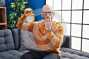 Young bald man with beard holding alarm clock covering mouth with hand, shocked and afraid for mistake