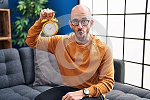 Young bald man with beard holding alarm clock scared and amazed with open mouth for surprise, disbelief face