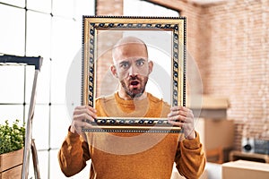 Young bald man with beard moving to a new home putting face inside vintage frame in shock face, looking skeptical and sarcastic,