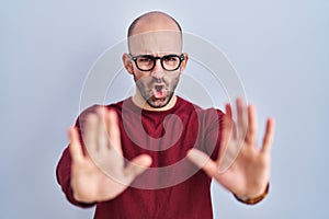 Young bald man with beard standing over white background wearing glasses doing stop gesture with hands palms, angry and