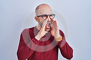 Young bald man with beard standing over white background wearing glasses shouting angry out loud with hands over mouth