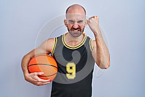 Young bald man with beard wearing basketball uniform holding ball angry and mad raising fist frustrated and furious while shouting