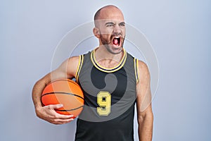Young bald man with beard wearing basketball uniform holding ball angry and mad screaming frustrated and furious, shouting with