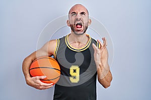 Young bald man with beard wearing basketball uniform holding ball crazy and mad shouting and yelling with aggressive expression