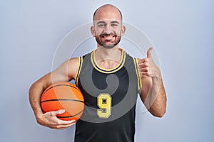Young bald man with beard wearing basketball uniform holding ball doing happy thumbs up gesture with hand