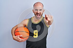 Young bald man with beard wearing basketball uniform holding ball pointing displeased and frustrated to the camera, angry and