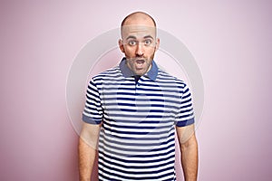 Young bald man with beard wearing casual striped blue t-shirt over pink isolated background afraid and shocked with surprise