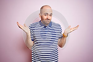 Young bald man with beard wearing casual striped blue t-shirt over pink isolated background clueless and confused expression with