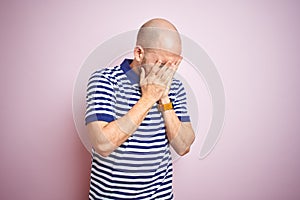 Young bald man with beard wearing casual striped blue t-shirt over pink isolated background with sad expression covering face with