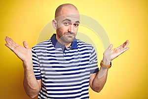 Young bald man with beard wearing casual striped blue t-shirt over yellow isolated background clueless and confused expression