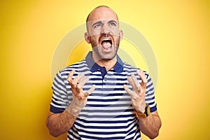 Young bald man with beard wearing casual striped blue t-shirt over yellow isolated background crazy and mad shouting and yelling