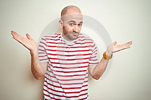 Young bald man with beard wearing casual striped red t-shirt over white isolated background clueless and confused expression with