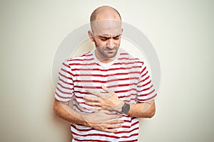 Young bald man with beard wearing casual striped red t-shirt over white isolated background with hand on stomach because