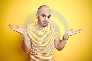 Young bald man with beard wearing casual striped t-shirt over yellow isolated background clueless and confused expression with