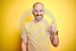 Young bald man with beard wearing casual striped t-shirt over yellow isolated background doing happy thumbs up gesture with hand
