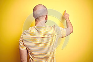 Young bald man with beard wearing casual striped t-shirt over yellow isolated background Posing backwards pointing ahead with