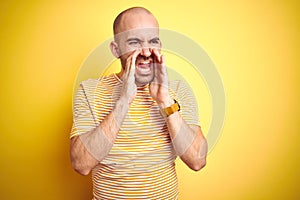 Young bald man with beard wearing casual striped t-shirt over yellow isolated background Shouting angry out loud with hands over