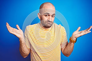 Young bald man with beard wearing casual t-shirt over blue isolated background clueless and confused expression with arms and