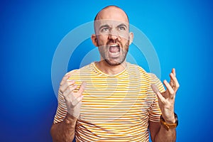 Young bald man with beard wearing casual t-shirt over blue isolated background crazy and mad shouting and yelling with aggressive